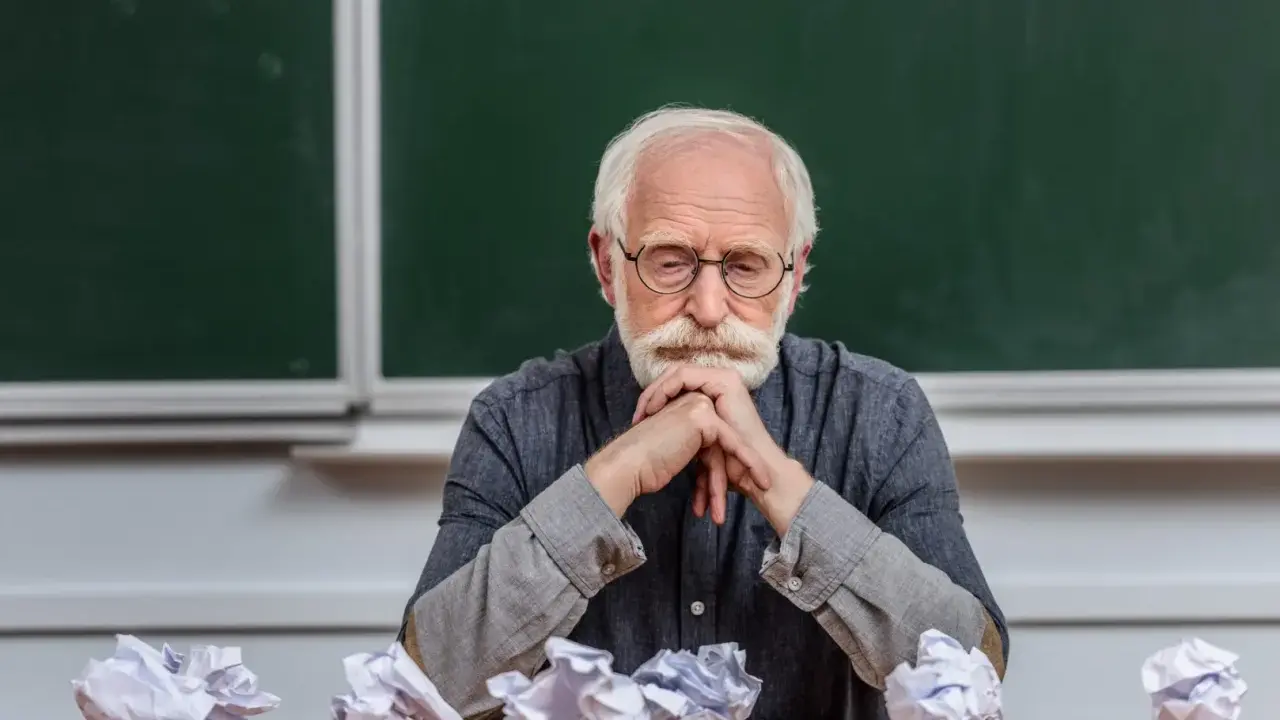 Zamyślony adiunkt, kto to? Siedzi przy biurku, przed nim pogięte kartki.