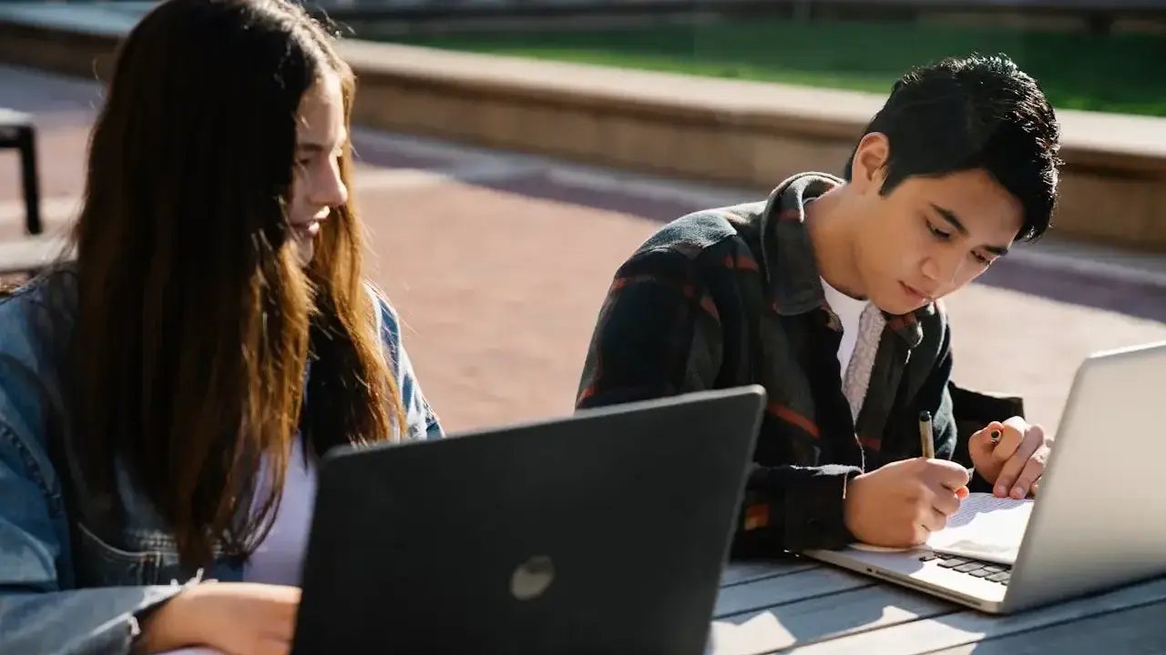 Dwoje studentów korzysta z laptopów na zewnątrz, przygotowując się do zajęć. To idealny przykład elastyczności, jaką oferują studia hybrydowe.