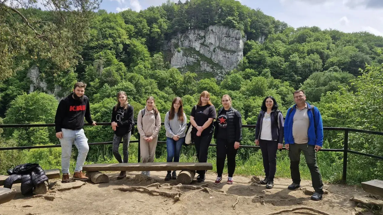 Grupa studentów studiów ochrona środowiska podziwia malowniczy krajobraz z klifem i bujną zielenią.