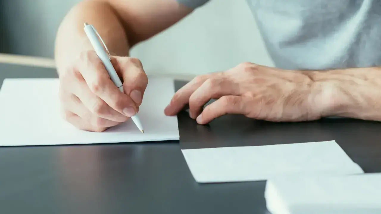 Ręce piszą list motywacyjny specjalista ds sprzedaży. Dokumenty i koperty leżą na biurku.