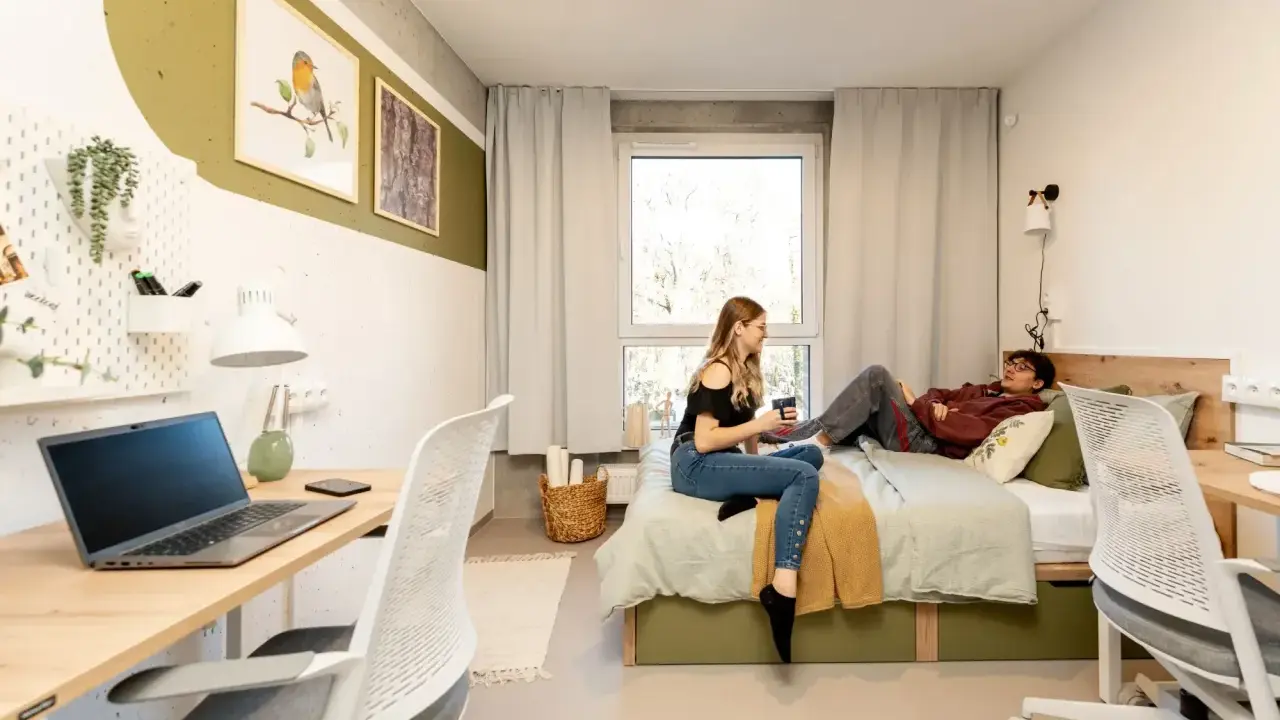 Dwie osoby w pokoju studenckim, jeden leży na łóżku, drugi siedzi obok. Tak wyglądają egzaminy na studiach – chwila wytchnienia przed nauką.