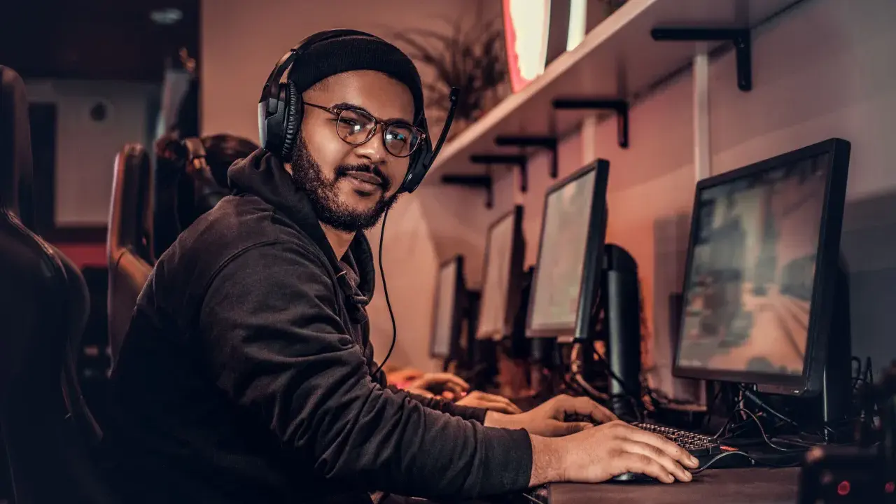 Młody game developer w słuchawkach i okularach pracuje przy komputerze w ciemnym pomieszczeniu.