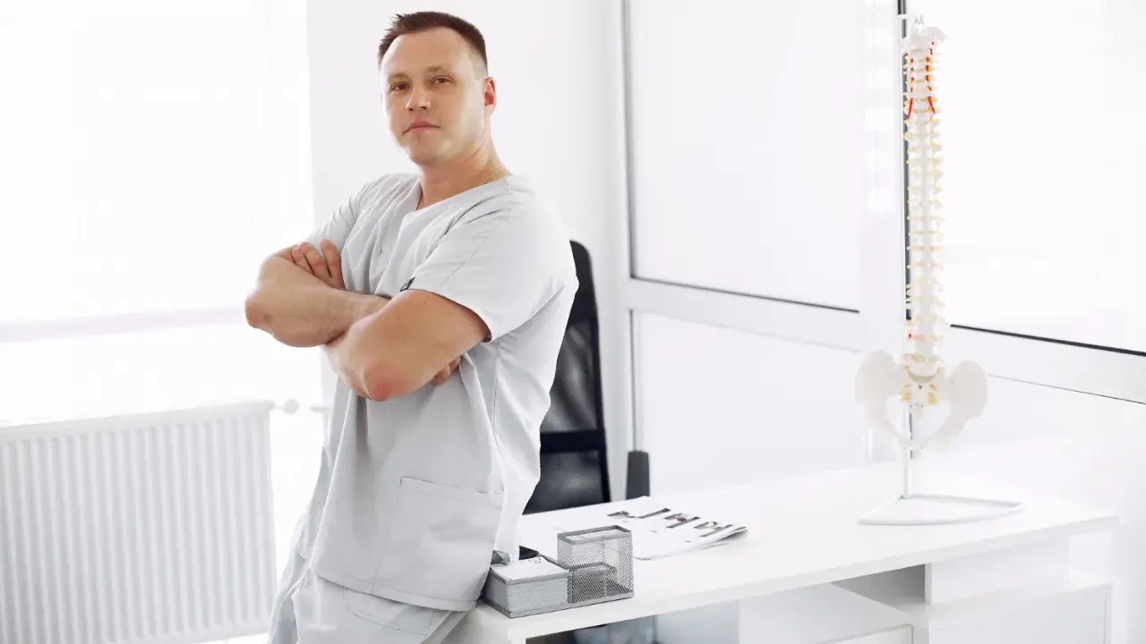 Mężczyzna w uniformie medycznej opiera się o biurko, na którym stoi model kręgosłupa. To przyszły absolwent wyższej szkoły rehabilitacji.