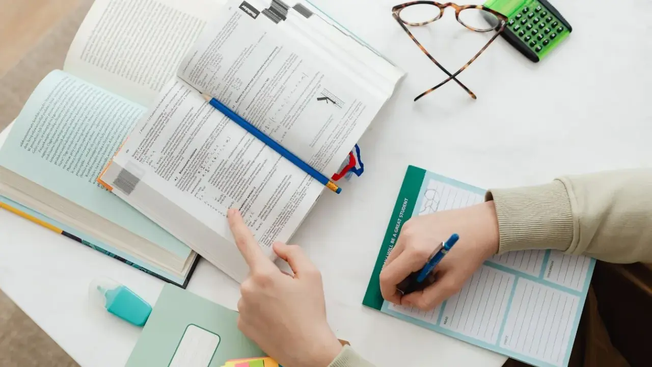 Ręce studenta nad otwartymi książkami, notatnik z napisem "I WILL BE A GREAT STUDENT", okulary i kalkulator. Nauka do egzaminu, by zdobyć tytuł zawodowy.