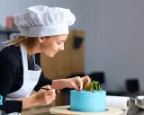 Uśmiechnięta cukierniczka w białym czepku i fartuchu dekoruje błękitny tort. Jej cv to pasja i precyzja w tworzeniu słodkich arcydzieł.