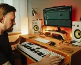 Mężczyzna tworzy muzykę w domowym studiu. Na biurku klawiatura sterująca, monitory studyjne, komputer z programem do produkcji muzyki i gitara.