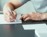 Ręce piszą list motywacyjny specjalista ds sprzedaży. Dokumenty i koperty leżą na biurku.