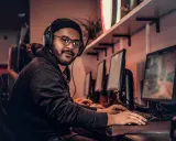 Młody game developer w słuchawkach i okularach pracuje przy komputerze w ciemnym pomieszczeniu.
