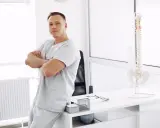 Mężczyzna w uniformie medycznej opiera się o biurko, na którym stoi model kręgosłupa. To przyszły absolwent wyższej szkoły rehabilitacji.