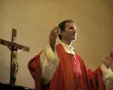 Kapłan w czerwonej szacie z podniesionymi rękami, obok krzyża z Jezusem. Wiernie przestrzega zasad prawa kanonicznego.