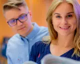 Uśmiechnięta studentka z blond włosami i dwoma kolegami w tle, dyskutujący nad materiałami. Być może przeglądają kozminski cennik.