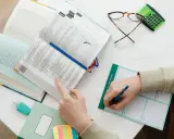 Ręce studenta nad otwartymi książkami, notatnik z napisem "I WILL BE A GREAT STUDENT", okulary i kalkulator. Nauka do egzaminu, by zdobyć tytuł zawodowy.