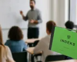 Zielony indeks na pierwszym planie, w tle sala wykładowa z nauczycielem i studentami. To symbol początku drogi do pracy po studiach.