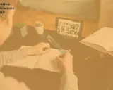 Osoba pisze w zeszycie, na stole tablet z wideokonferencją i otwarta książka.