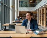 Młody mężczyzna zamyślony nad książkami i laptopem w bibliotece. Myśli o przeniesieniu się na inną uczelnię.