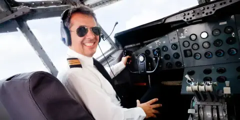 Uśmiechnięty pilot w okularach przeciwsłonecznych i słuchawkach siedzi w kokpicie. Zastanawiasz się, ile zarabia pilot samolotu?