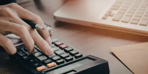 Osoba liczy na kalkulatorze, zastanawiając się, czy studia stacjonarne są płatne. Obok leży laptop i koperta.
