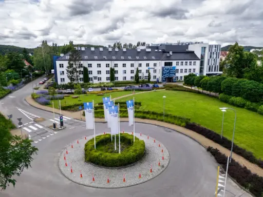 Nowoczesny budynek WSB Gdańsk otoczony zielenią. Na rondzie powiewają białe flagi z logo uczelni.