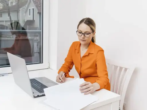 Kobieta w okularach i pomarańczowej bluzce analizuje dokumenty przy laptopie. Dowiaduje się, jak zostać notariuszem.