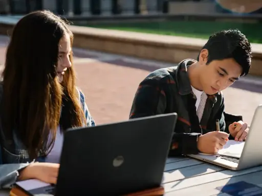 Dwoje studentów korzysta z laptopów na zewnątrz, przygotowując się do zajęć. To idealny przykład elastyczności, jaką oferują studia hybrydowe.