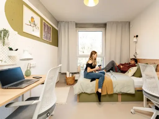 Dwie osoby w pokoju studenckim, jeden leży na łóżku, drugi siedzi obok. Tak wyglądają egzaminy na studiach – chwila wytchnienia przed nauką.