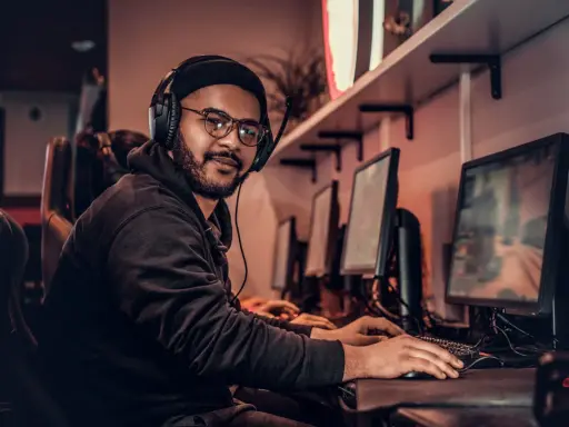 Młody game developer w słuchawkach i okularach pracuje przy komputerze w ciemnym pomieszczeniu.