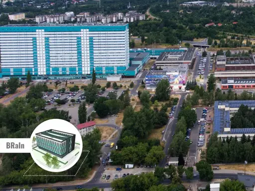 Widok z lotu ptaka na kompleks budynków, w tym duży, jasny budynek z niebieskimi akcentami, przypominający umed w Łodzi. Widać parkingi, drogi i zieleń.