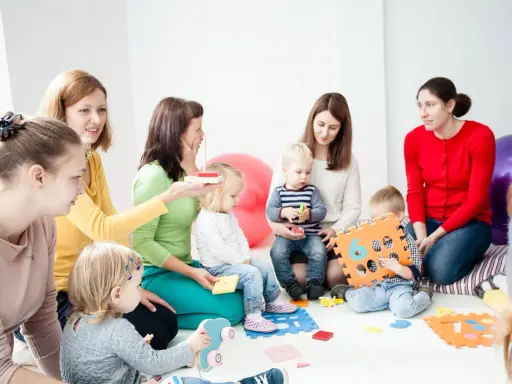 Grupa dzieci i dorosłych uczestniczy w zajęciach, które przypominają terapię pedagogiczną. Dzieci bawią się klockami i zabawkami.