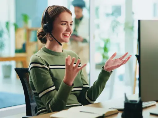Uśmiechnięta customer experience manager w słuchawkach gestykuluje, siedząc przy biurku przed monitorem.