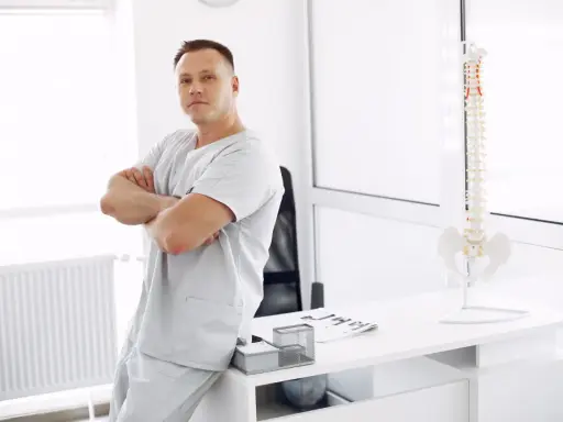 Mężczyzna w uniformie medycznej opiera się o biurko, na którym stoi model kręgosłupa. To przyszły absolwent wyższej szkoły rehabilitacji.