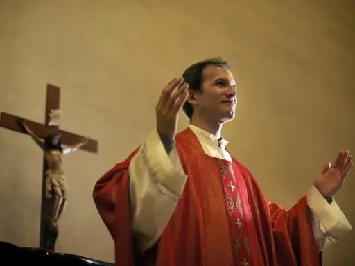Kapłan w czerwonej szacie z podniesionymi rękami, obok krzyża z Jezusem. Wiernie przestrzega zasad prawa kanonicznego.
