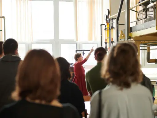 Grupa studentów na zajęciach praktycznych. Kobieta w czerwonej sukience tłumaczy coś przy aparaturze. To może być część ich studiów magisterskich zaocznych w Warszawie.