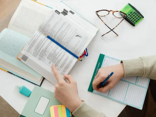 Ręce studenta nad otwartymi książkami, notatnik z napisem "I WILL BE A GREAT STUDENT", okulary i kalkulator. Nauka do egzaminu, by zdobyć tytuł zawodowy.