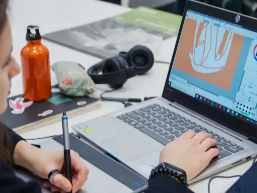 Studentka pracuje nad projektem w **studiach graficznych Warszawa**. Na ekranie laptopa widać program do grafiki, a obok słuchawki i butelkę z wodą.