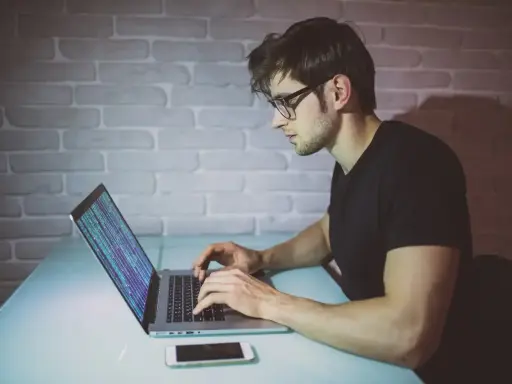 Młody mężczyzna w okularach pisze kod na laptopie. To jego pierwszy krok, by dowiedzieć się, jak zostać informatykiem.