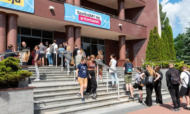 Grupa młodych ludzi schodzi po schodach przed budynkiem, gdzie wisi baner "Spotkajmy się w Kortowie!". To idealne miejsce na rozpoczęcie studiów w Olsztynie.
