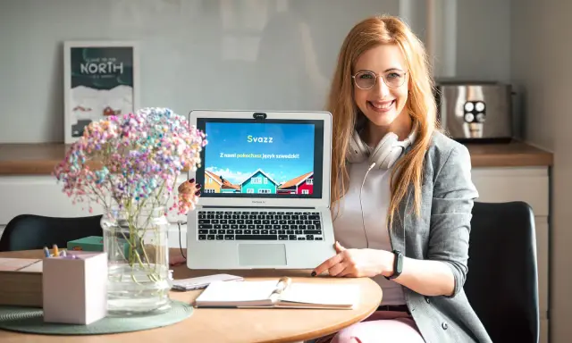 Uśmiechnięta kobieta prezentuje laptop z napisem "Z nami pokochasz język szwedzki!". Idealna dla studentów filologii szwedzkiej w Warszawie.