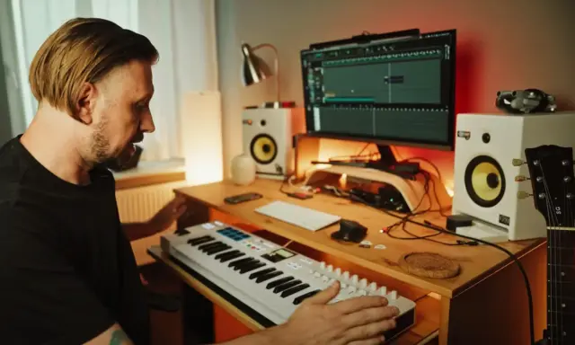 Mężczyzna tworzy muzykę w domowym studiu. Na biurku klawiatura sterująca, monitory studyjne, komputer z programem do produkcji muzyki i gitara.