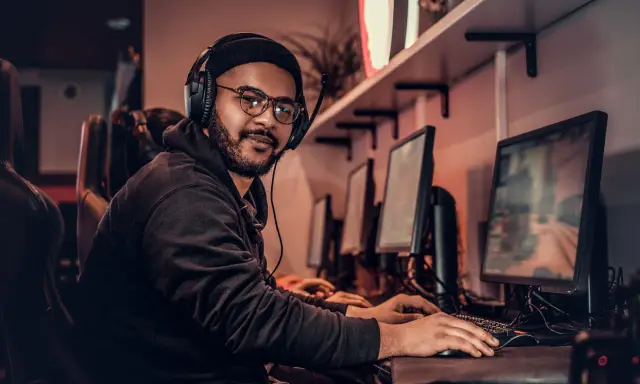 Młody game developer w słuchawkach i okularach pracuje przy komputerze w ciemnym pomieszczeniu.