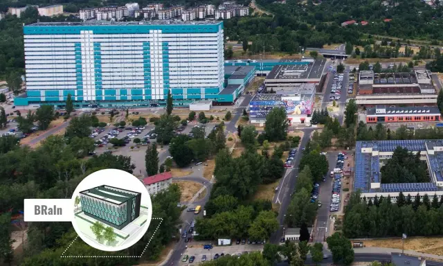 Widok z lotu ptaka na kompleks budynków, w tym duży, jasny budynek z niebieskimi akcentami, przypominający umed w Łodzi. Widać parkingi, drogi i zieleń.