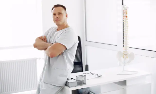 Mężczyzna w uniformie medycznej opiera się o biurko, na którym stoi model kręgosłupa. To przyszły absolwent wyższej szkoły rehabilitacji.