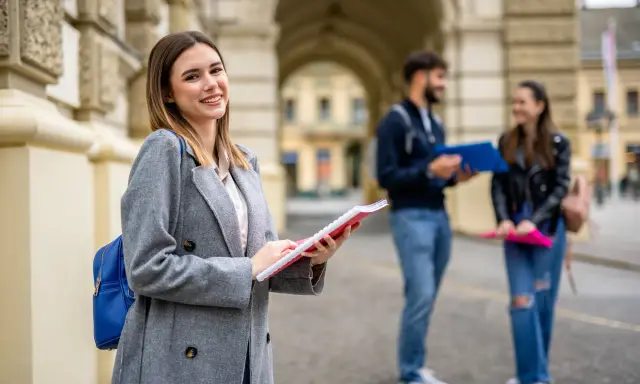 Uśmiechnięta studentka z notatnikiem w ręku, przygotowująca się do studiów 2 stopnia. W tle para studentów.