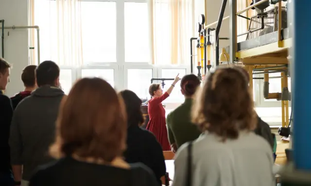 Grupa studentów na zajęciach praktycznych. Kobieta w czerwonej sukience tłumaczy coś przy aparaturze. To może być część ich studiów magisterskich zaocznych w Warszawie.