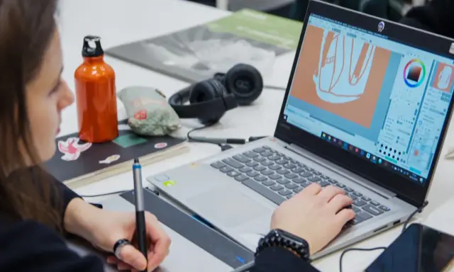 Studentka pracuje nad projektem w **studiach graficznych Warszawa**. Na ekranie laptopa widać program do grafiki, a obok słuchawki i butelkę z wodą.