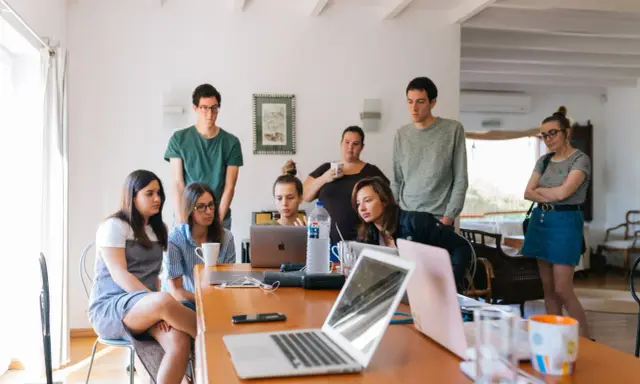 Grupa studentów na którym roku studiów są praktyki, pochylona nad laptopami, dyskutuje przy drewnianym stole.