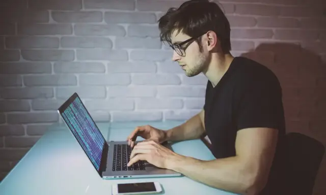 Młody mężczyzna w okularach pisze kod na laptopie. To jego pierwszy krok, by dowiedzieć się, jak zostać informatykiem.
