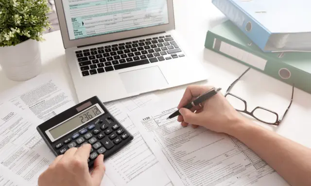 Osoba wypełnia formularz podatkowy, korzystając z kalkulatora i laptopa. To może być początek drogi, jak zostać księgowym.