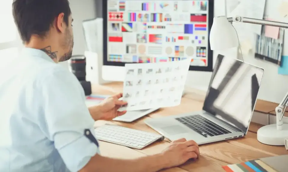 UX UI designer pracuje przy biurku, analizując wydruki i ekran komputera z paletą barw.