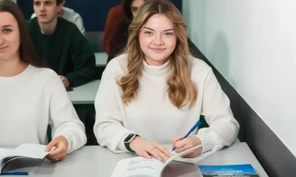 Uśmiechnięta studentka pisze notatki, zastanawiając się, ile wynosi stypendium na studiach. W tle inni studenci.