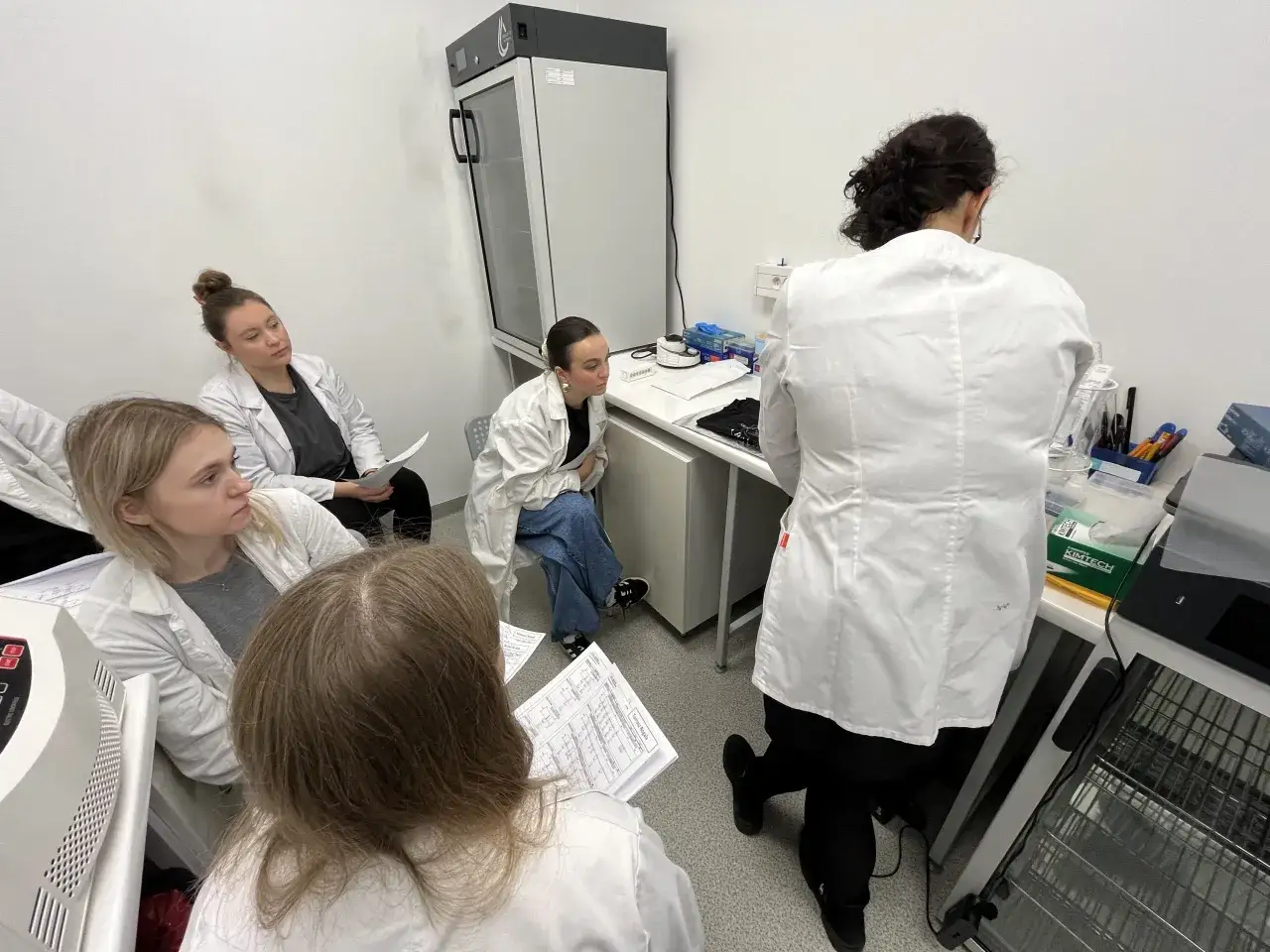 Studenci w kitlach laboratoryjnych słuchają wykładu z biologia kryminalistyczna, analizując dokumenty.